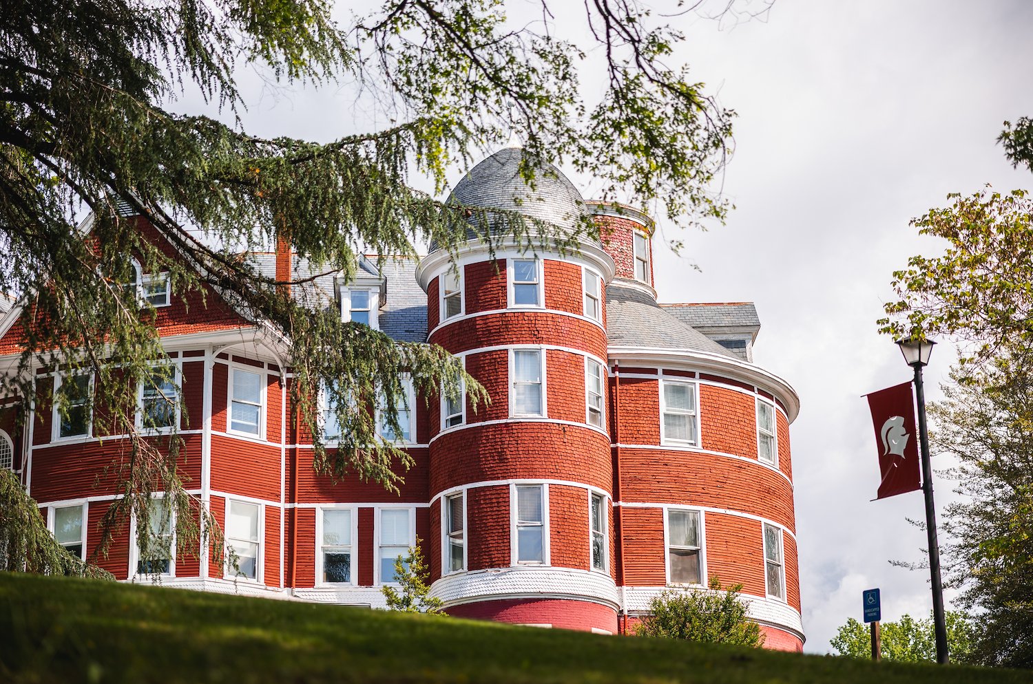 A vibrant red, multi-story building with white trim and numerous windows, featuring a prominent rounded turret with a conical roof on the right side. The building is partially obscured by green tree branches in the foreground and on the left. A dark red banner with a white emblem hangs from a lamppost on the far right. The sky is cloudy.