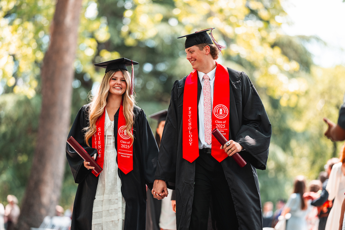 A couple dressed in graduation robes hold hands