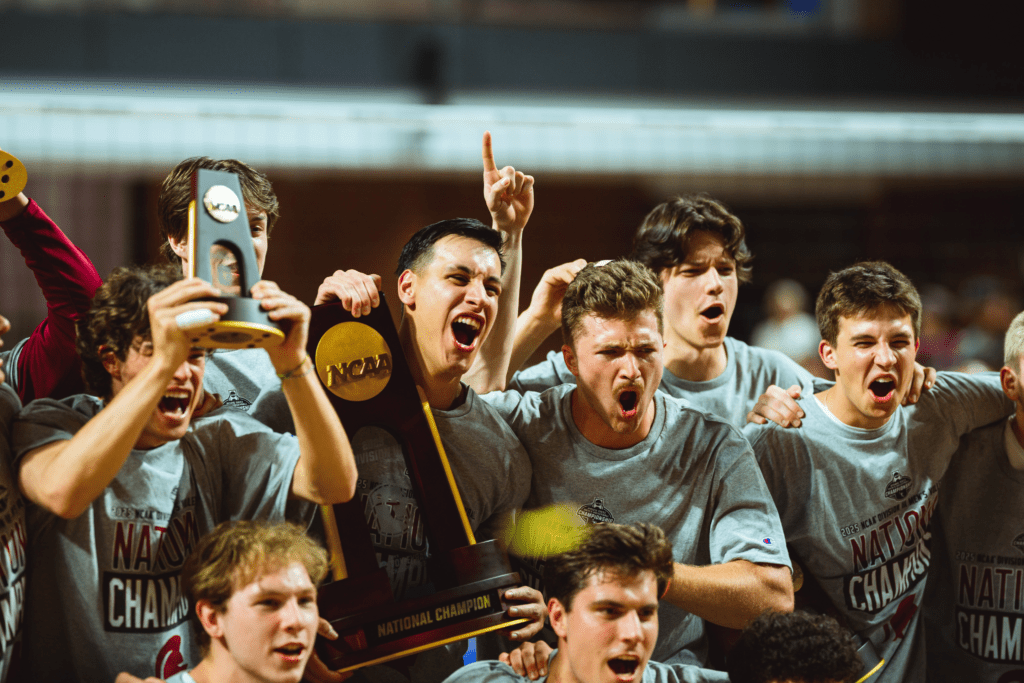 A jubilant group of young male athletes celebrating a national championship. They are wearing matching gray shirts, holding NCAA trophies, and cheering joyfully.