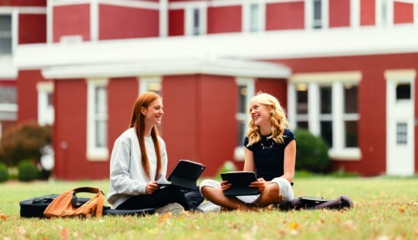 Two young women sit on grass outside a red building, smiling and holding tablets. They appear relaxed and engaged, conveying a mood of friendship and study.