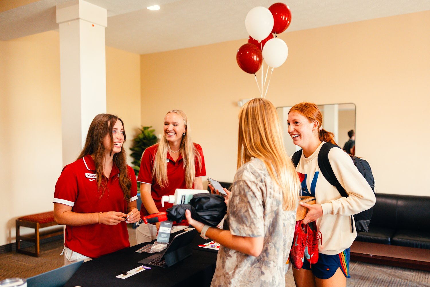 Two smiling admissions staff members in red shirts greeting prospective students at a check-in desk with red and white balloons.